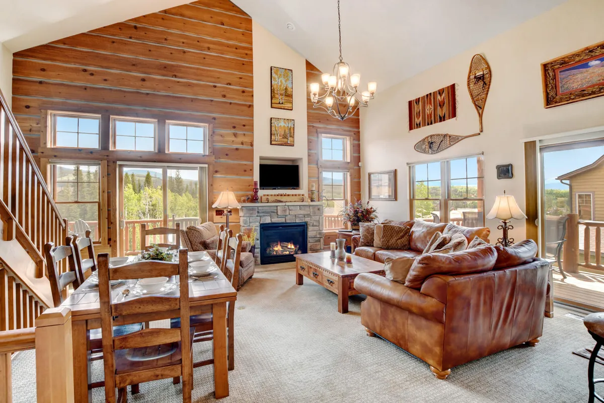 A warm, inviting living and dining area with a large stone fireplace, expansive windows with mountain views, a substantial dining table, plush leather sofas, and a wooden staircase, all bathed in natural light.