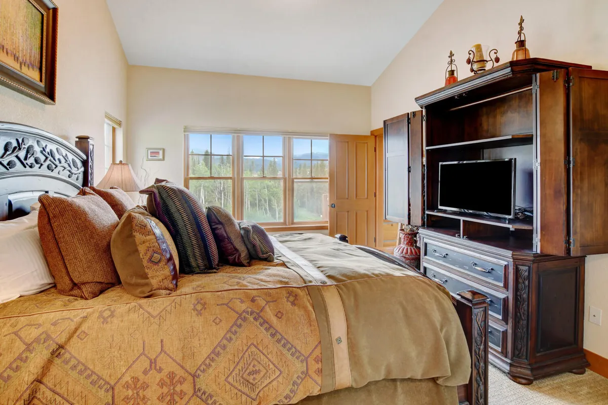 A cozy bedroom featuring a bed with decorative pillows, a wooden armoire with a built-in TV, and large windows that let in natural light. The space feels both inviting and comfortable, perfect for relaxation or unwinding with a favorite show.