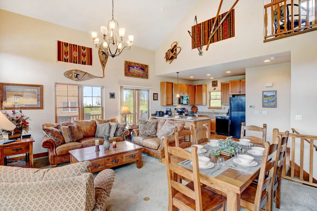 An open-plan space combining a comfortable seating area, a dining table set for a meal, and visible kitchen appliances. Decorative wall art enhances the homey feel, suggesting a space ideal for group meals and lively conversation.