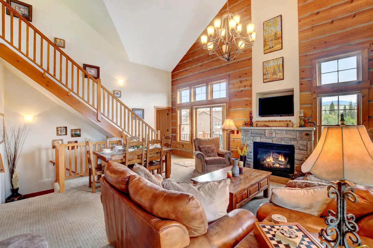 An inviting interior scene centered around a fireplace, with a large dining table, elegant chandelier, broad windows bringing in mountain light, and a visible staircase, all styled in a welcoming, cozy manner.