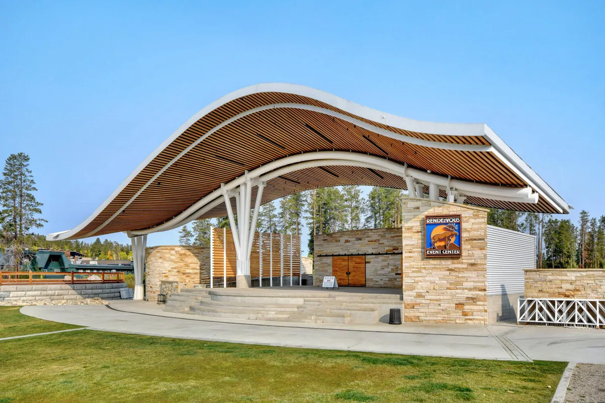 An exterior shot of a modern amphitheater with a distinctive curved wooden roof and stone façade, enhanced by clear event signage. The space is designed for live performances, community gatherings, and festivals, evoking a sense of excitement and shared experience.