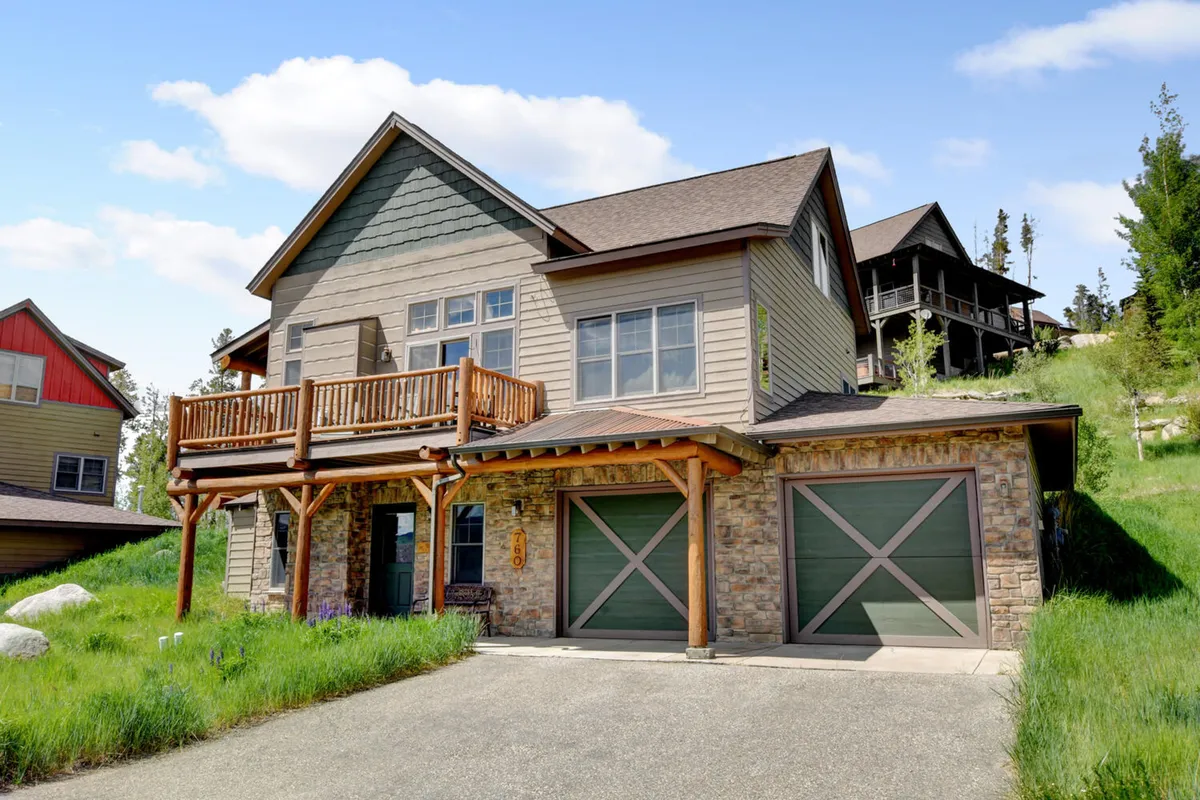 The hero image showcases the exterior of a luxurious mountain cabin with a striking stone facade and inviting wooden balcony. Lush green grass surrounds the home, and spacious garage doors hint at ample parking. The scene is inviting and relaxed, with hints of family gatherings and outdoor fun. The balcony provides an ideal spot to enjoy scenic mountain views, morning coffee, and evening barbecues under a colorful sunset sky.