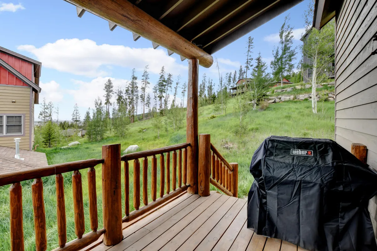 An exterior shot of a deck with a wooden railing and a grill covered with a black cover. Lush green grass and distant trees set a peaceful, private outdoor scene, ideal for al fresco dining or grilling.