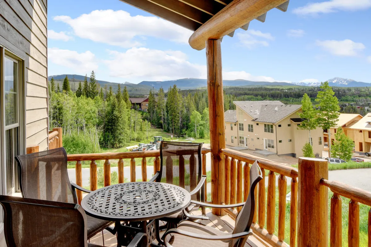 A circular table with chairs sits on a wooden balcony, overlooking a breathtaking panorama of forested mountains and blue sky. The setting is tranquil, perfect for enjoying morning coffee or evening relaxation amidst nature.