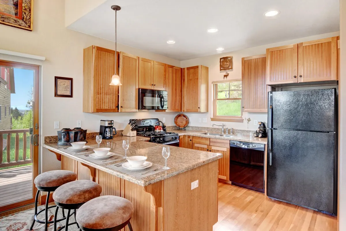 A well-lit, inviting kitchen featuring a granite countertop, warm wood cabinetry, modern kitchen appliances, and stools, with a window offering a scenic view. The setting is designed for both culinary creativity and casual conversation.