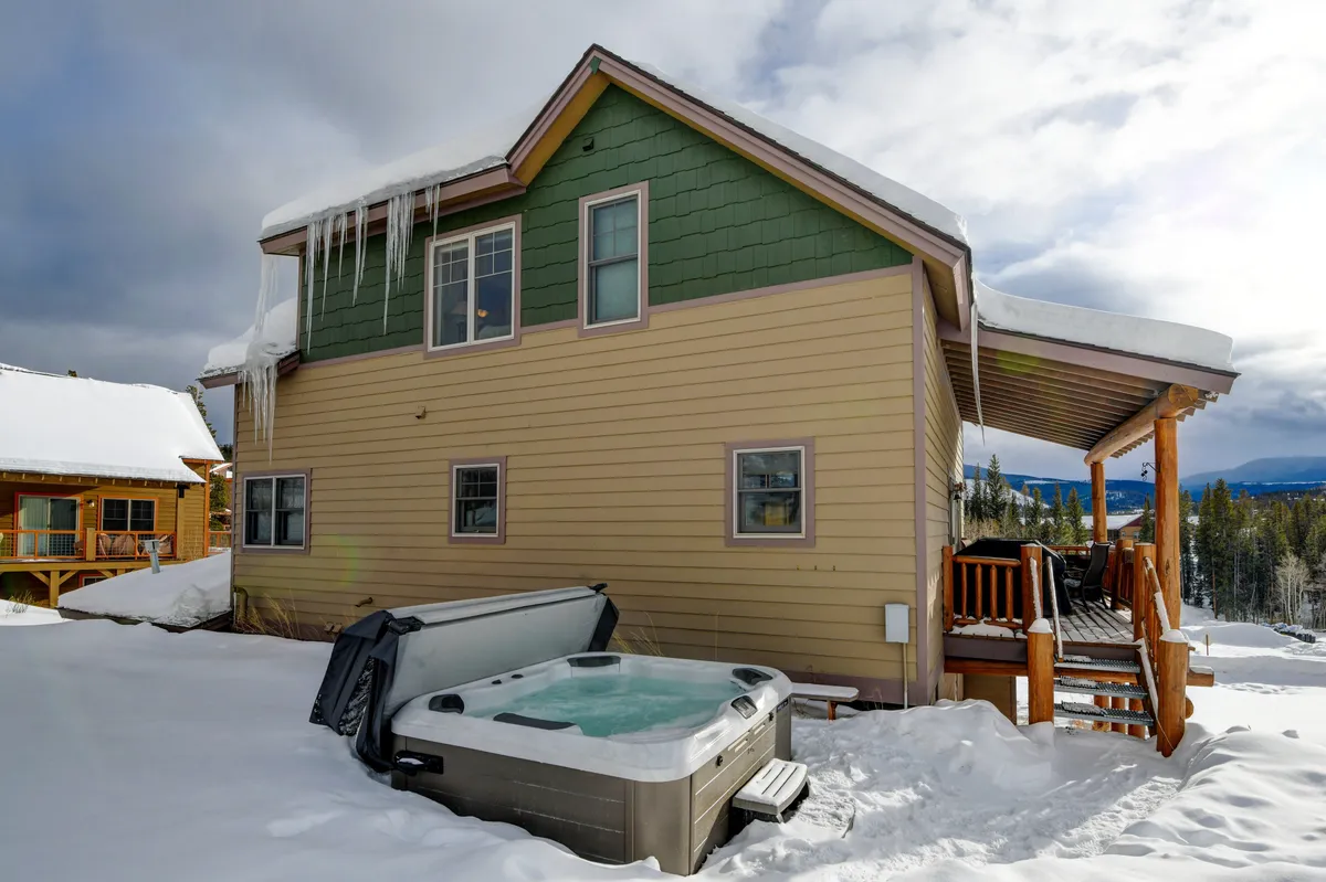 A steaming hot tub sits on a spacious wooden deck, surrounded by a winter wonderland with snow-covered ground and icicles hanging from the roof above. Beyond the deck, pine trees and mountain scenery create a peaceful alpine backdrop. The scene exudes relaxation and indulgence amid the elements.