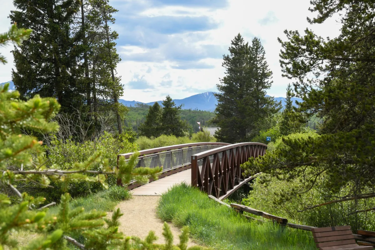 A rustic wooden bridge blends into lush greenery with a dramatic mountain backdrop. The path invites exploration, suggesting access to neighborhood trails and opportunities to step outside for wellness breaks or midday walks.