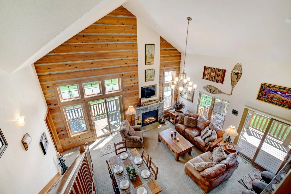 A spacious great room featuring a soaring vaulted ceiling, large windows showcasing stunning mountain views, a stone fireplace at the center, comfortable seating, and a dining table set for gatherings.