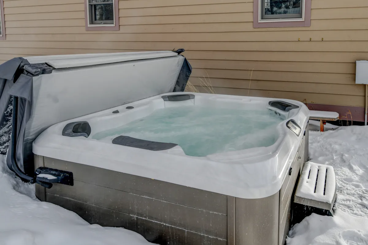 An inviting outdoor hot tub sits on a snow-covered patio, surrounded by nature and a wooden bench. The scene captures the steam rising from the warm water, offering a sense of tranquility and luxury while hinting at the wintry adventure just beyond.