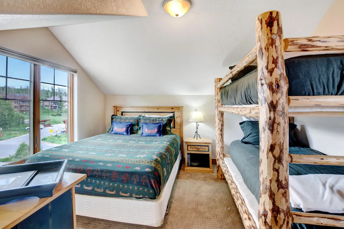 A welcoming guest room that combines a bunk bed with a queen bed, illuminated by a large window. A nightstand with a lamp provides a homey touch, making it suitable for kids, friends, or families.