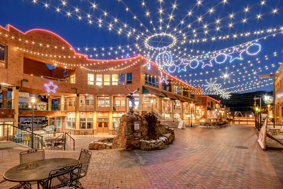The exterior evening scene features string lights suspended overhead, decorative stars, a central fountain sculpture, and a welcoming outdoor seating area. The setting is lively and festive, suggesting a place where friends and groups can gather, dine, and enjoy community activities after dark.