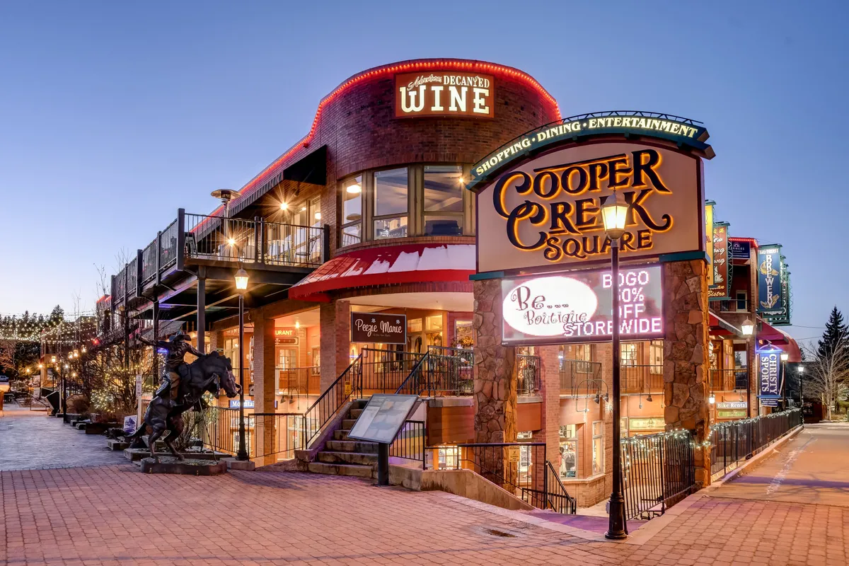 The vibrant exterior of the Cooper Creek Square shopping and dining complex is featured, with a welcoming sign, a wine shop, outdoor seating, and decorative lighting. The scene bustles with activity, offering a glimpse into the lively local area and its amenities.
